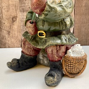 May include: A green and brown gnome figurine with a red hat and a basket of fish. The gnome is sitting on a rock and holding a fishing rod.