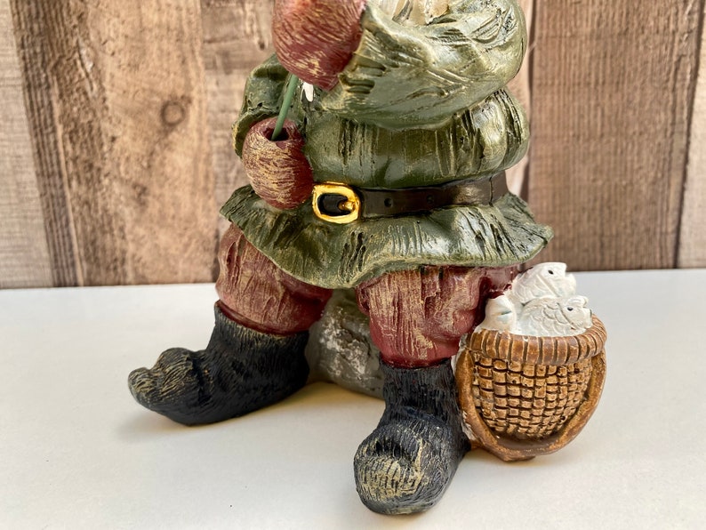 May include: A green and brown gnome figurine with a red hat and a basket of fish. The gnome is sitting on a rock and holding a fishing rod.