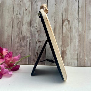 May include: A white picture frame with a gold-colored decorative top. The frame has a black stand that folds out to prop it up.