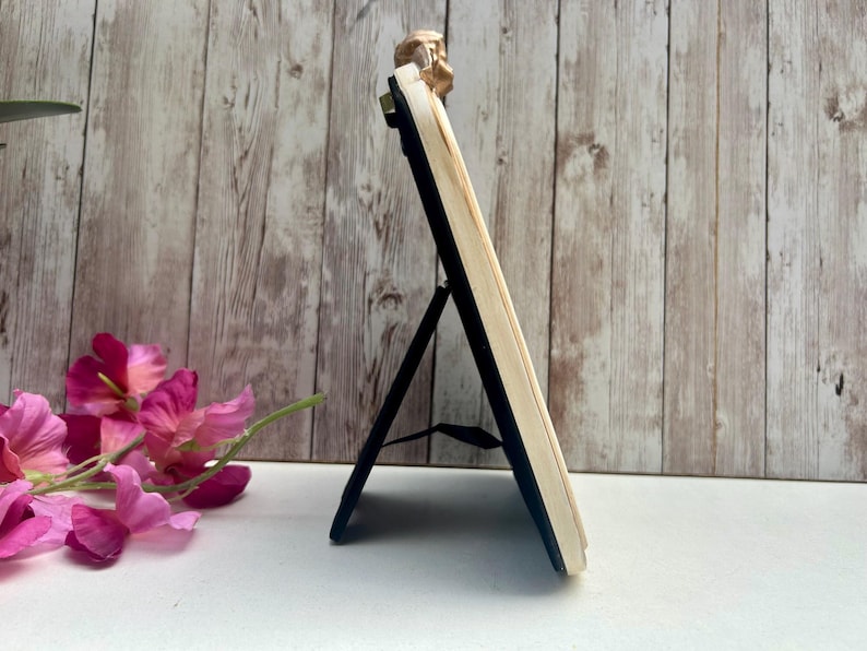 May include: A white picture frame with a gold-colored decorative top. The frame has a black stand that folds out to prop it up.