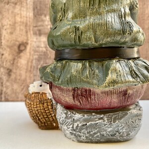 May include: A green and red gnome figurine with a basket in its hands. The gnome is wearing a green hat and a brown belt. The gnome is sitting on a stone base.