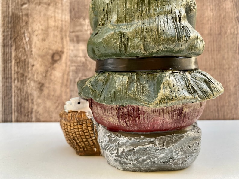 May include: A green and red gnome figurine with a basket in its hands. The gnome is wearing a green hat and a brown belt. The gnome is sitting on a stone base.