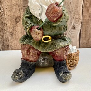 May include: A green and brown gnome figurine sitting on a rock with a fishing rod in its hand. The gnome is wearing a green hat and a brown belt. The gnome is holding a basket of white flowers.