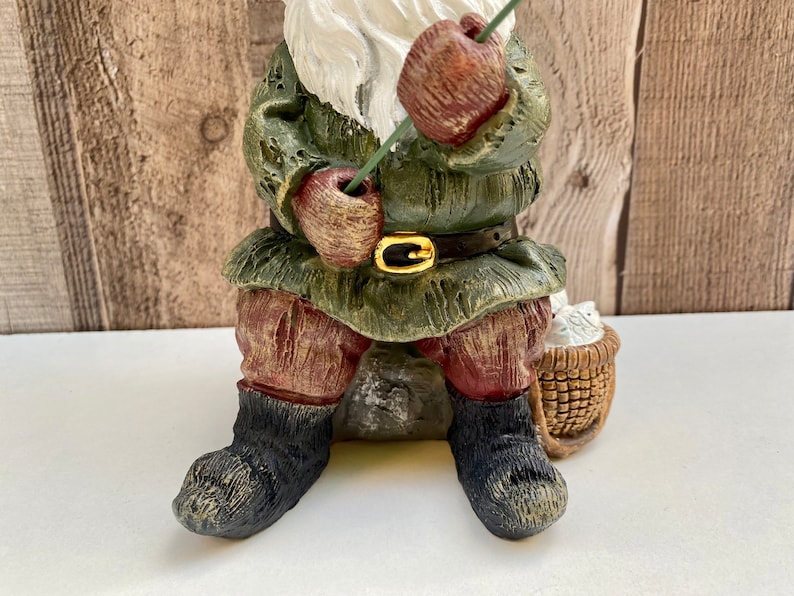 May include: A green and brown gnome figurine sitting on a rock with a fishing rod in its hand. The gnome is wearing a green hat and a brown belt. The gnome is holding a basket of white flowers.