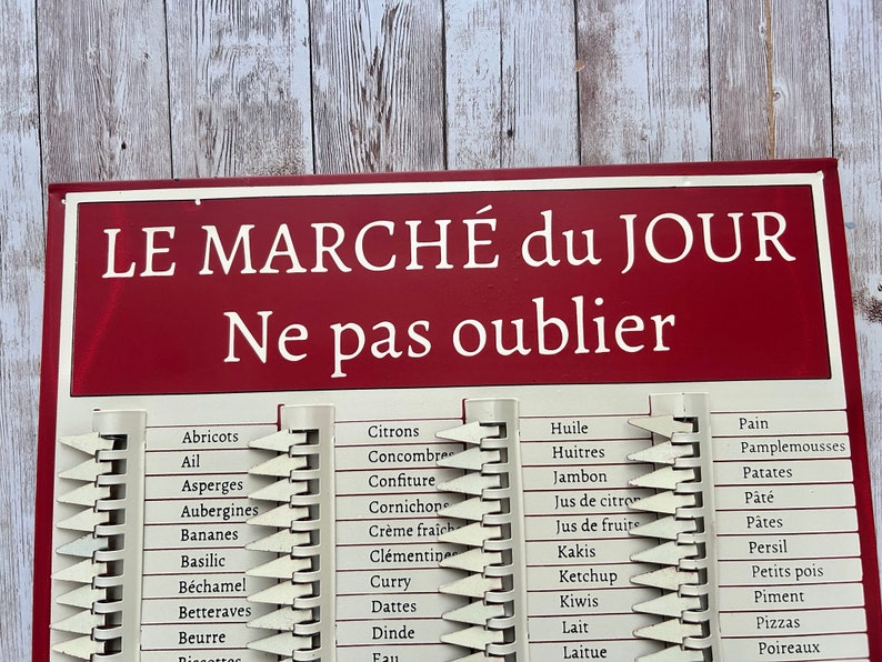 Op de afbeelding: Een rood en wit bord met de tekst "LE MARCH&Eacute; du JOUR Ne pas oublier" in wit. Het bord bevat een lijst met voedingsmiddelen in het Frans, zoals "Abricots", "Citrons", "Huile" en "Pain".