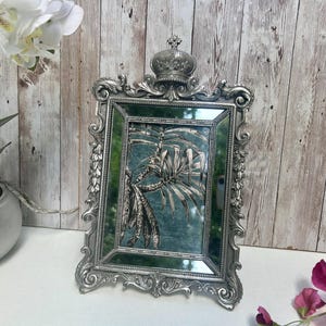 May include: Silver ornate picture frame with a green mirrored background and a crown on top. The frame is sitting on a white surface.
