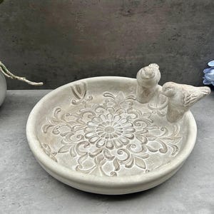 May include: A white ceramic bird bath with a decorative floral design and two small bird figurines. The bird bath is ideal for attracting birds to your garden.