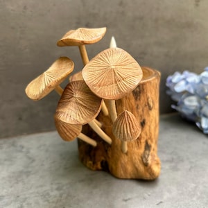 May include: A wooden mushroom sculpture featuring carved fungi sprouting from a hollowed log. The mushrooms have detailed caps and stems, and the wood has a natural, warm tone. A small white candle is visible.