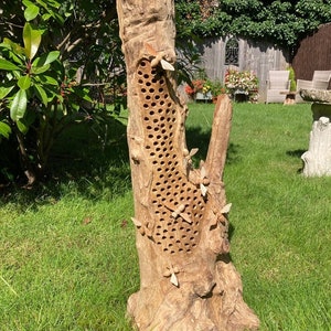 K&ouml;nnte beinhalten: Ein Bienenhaus aus Holz in Form eines Baumstamms mit einem Wabenmuster und geschnitzten Bienen, die umherfliegen. Das Bienenhaus steht auf einem Bett aus gr&uuml;nem Gras.