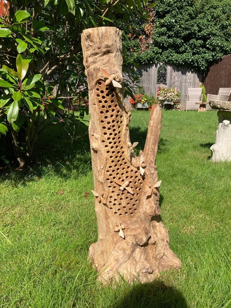 K&ouml;nnte beinhalten: Ein Bienenhaus aus Holz in Form eines Baumstamms mit einem Wabenmuster und geschnitzten Bienen, die umherfliegen. Das Bienenhaus steht auf einem Bett aus gr&uuml;nem Gras.