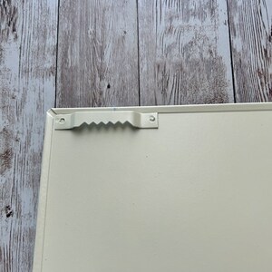Op de afbeelding: De achterkant van een wit metalen bord met een handvat. Het handvat is van metaal en heeft een golvend ontwerp.