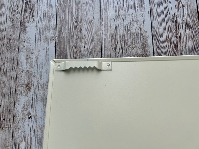 Op de afbeelding: De achterkant van een wit metalen bord met een handvat. Het handvat is van metaal en heeft een golvend ontwerp.