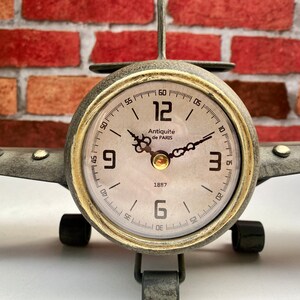 May include: A vintage-style aeroplane clock with a gold-tone finish and a white face. The clock features black Roman numeral hour markers and black hands. The clock face also includes the text "Antiquit&eacute; de PARIS" and "1887".