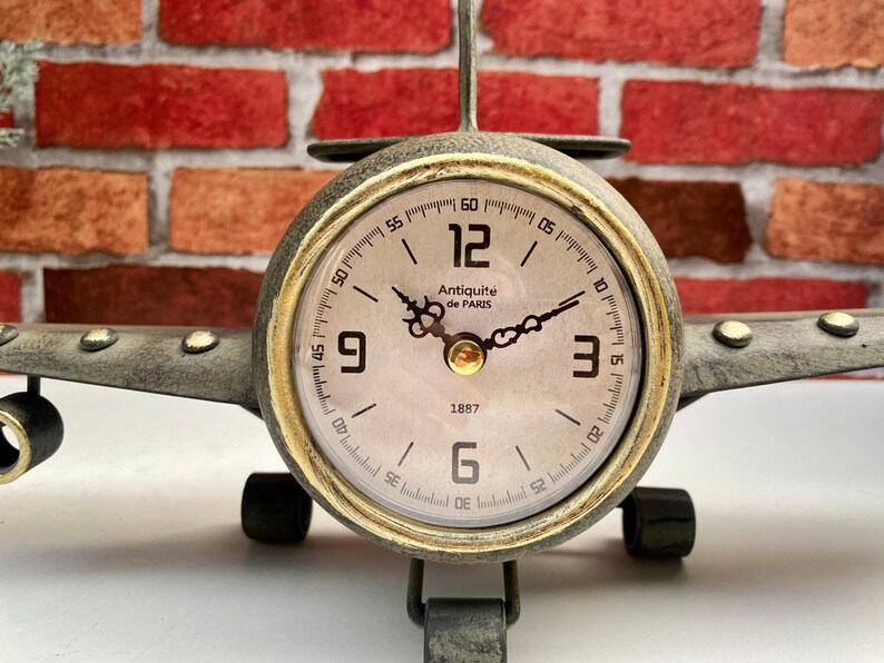 May include: A vintage-style aeroplane clock with a gold-tone finish and a white face. The clock features black Roman numeral hour markers and black hands. The clock face also includes the text "Antiquit&eacute; de PARIS" and "1887".