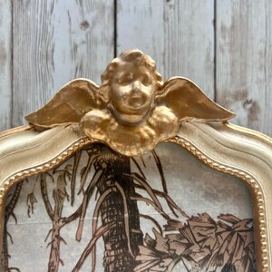 May include: A gold-painted picture frame with a cherub design. The frame is white with gold accents and has a decorative border. The frame is empty and ready for a picture.