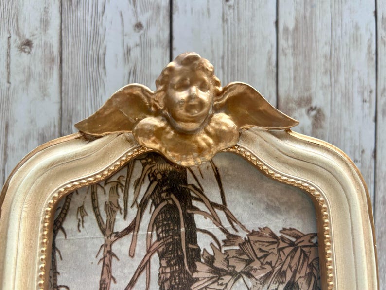 May include: A gold-painted picture frame with a cherub design. The frame is white with gold accents and has a decorative border. The frame is empty and ready for a picture.