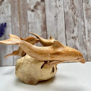 Peut inclure: Une sculpture en bois repr&eacute;sentant trois dauphins sautant d'une base de couleur claire. Les dauphins sont sculpt&eacute;s dans du bois brun clair, avec des veines visibles et des nageoires d&eacute;taill&eacute;es. La sculpture est pos&eacute;e sur un fond en panneaux de bois.