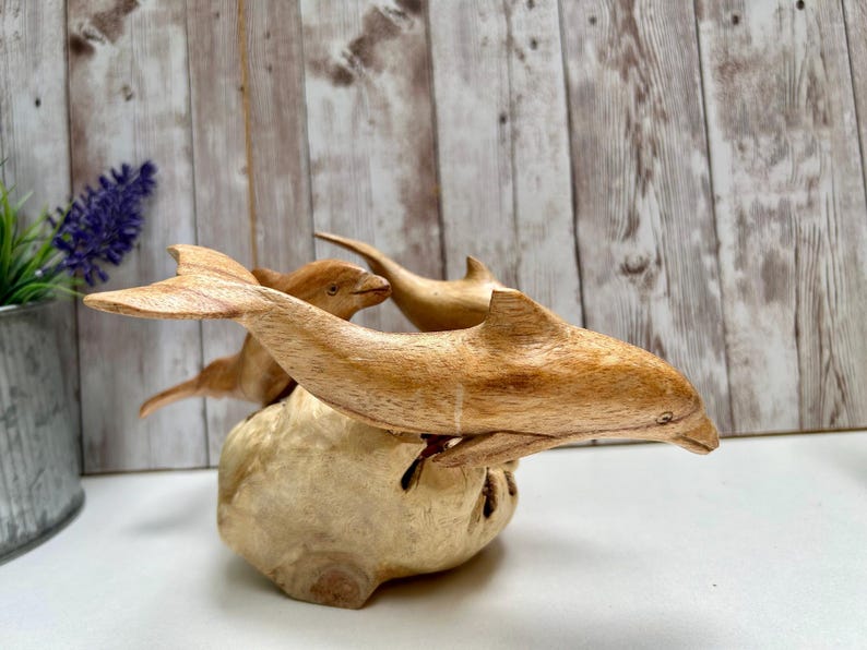 Peut inclure: Une sculpture en bois repr&eacute;sentant trois dauphins sautant d'une base de couleur claire. Les dauphins sont sculpt&eacute;s dans du bois brun clair, avec des veines visibles et des nageoires d&eacute;taill&eacute;es. La sculpture est pos&eacute;e sur un fond en panneaux de bois.