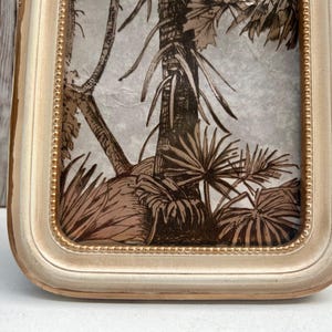 May include: A white frame with a gold trim surrounds a print of a tropical leaf pattern. The print is in shades of brown and beige.