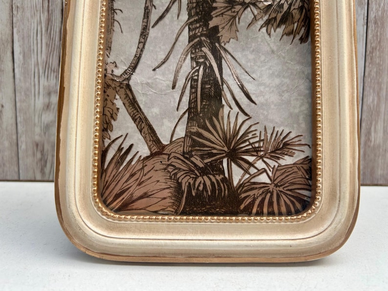 May include: A white frame with a gold trim surrounds a print of a tropical leaf pattern. The print is in shades of brown and beige.
