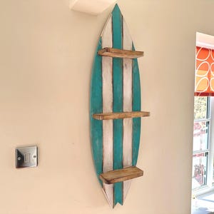 May include: A vertical wall-mounted shelf shaped like a surfboard, featuring alternating vertical stripes of teal and white paint. The unit includes three horizontal wooden shelves with a natural brown finish, evenly spaced along the centre.