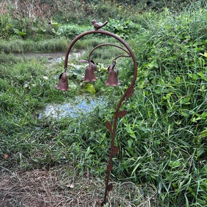 May include: A brown metal garden stake featuring a curved stem with small leaves, topped with a small bird figure. Three bell-shaped flower ornaments hang from the curved top section, each suspended by a thin wire, creating a decorative structure.