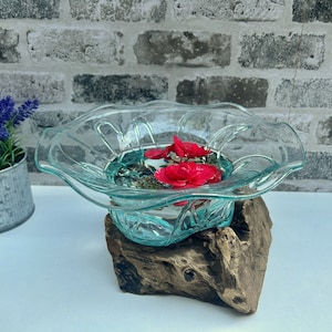 May include: A clear glass bowl with a scalloped edge, containing red flowers in water, placed on driftwood. The bowl is filled with water and sits on a wooden base. A small metal bucket with purple flowers is in the background.