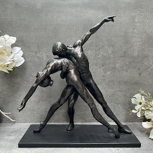May include: Bronze sculpture depicting two figures in a dance pose. One figure is arched backward, supported by another figure reaching upwards. The sculpture is mounted on a black rectangular base. White flowers are in the background.