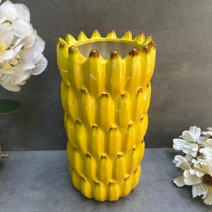 May include: A cylindrical ceramic vase featuring a bright yellow, three-dimensional relief pattern of stacked bananas. The top rim has a scalloped edge formed by the banana tips, which are accented with brown detailing. The interior is smooth and white.