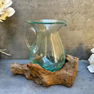 Peut inclure: Une carafe en verre transparent avec une anse et un bec, posée sur un morceau de bois flotté. La carafe a une base et un bord de couleur turquoise. Le fond présente des fleurs blanches et une surface grise.