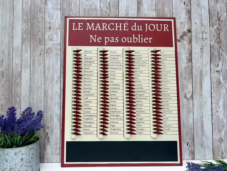 Op de afbeelding: Een rood en wit metalen boodschappenlijstbord met de tekst "LE MARCH&Eacute; du JOUR Ne pas oublier" bovenaan. Het bord heeft een lijst met Franse voedingsmiddelen in kolommen, waarbij elk item op een aparte regel staat. Het bord heeft een zwart schoolbord onderaan.