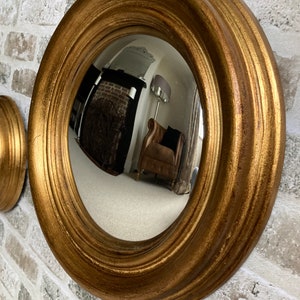May include: A gold-toned round mirror with a convex lens, framed with a thick, ornate, and slightly raised border. The mirror is mounted on a brick wall.