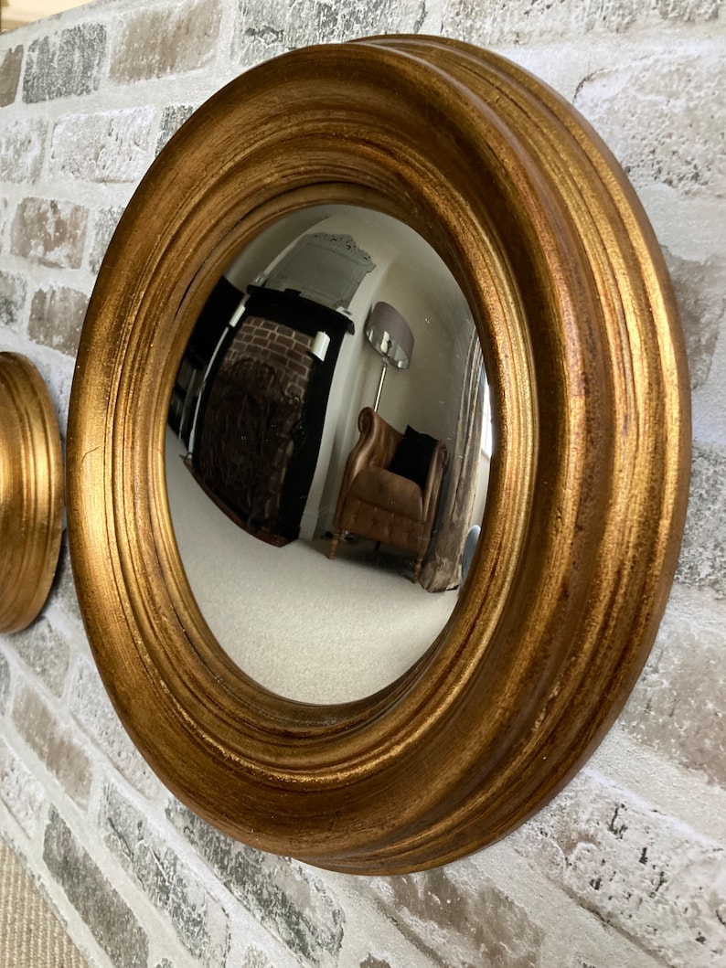 May include: A gold-toned round mirror with a convex lens, framed with a thick, ornate, and slightly raised border. The mirror is mounted on a brick wall.