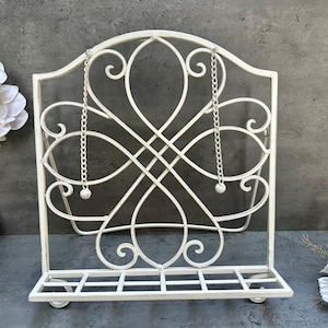 May include: A white metal decorative stand with an ornate, scrolled design. The stand features a shelf at the bottom and is suspended by chains. The stand is designed to hold books or display items. The stand is painted white.