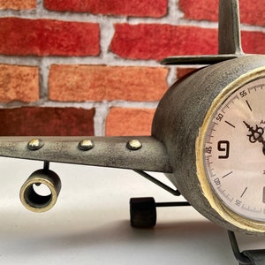 May include: A vintage airplane clock with a grey metal body and a gold-rimmed clock face. The clock face has black Roman numeral hour markers and black hands. The clock face also has the text "Antiquit&eacute; de PARIS" and "1887".