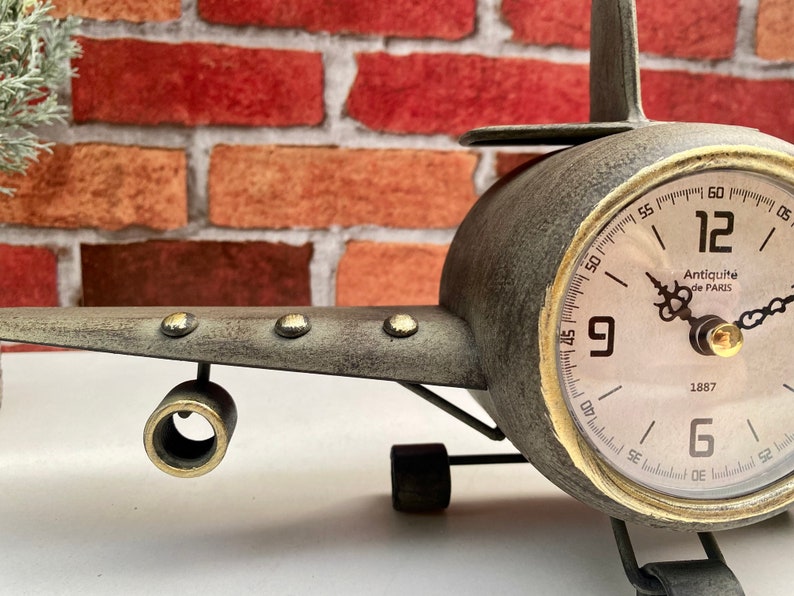 May include: A vintage airplane clock with a grey metal body and a gold-rimmed clock face. The clock face has black Roman numeral hour markers and black hands. The clock face also has the text "Antiquit&eacute; de PARIS" and "1887".