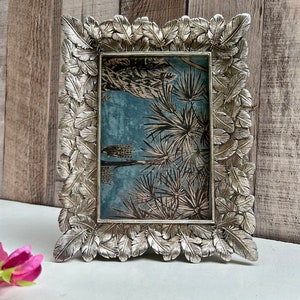 May include: Silver picture frame with a leafy design. The frame is empty and has a light blue background with a floral pattern.