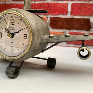 May include: A grey metal aeroplane clock with gold accents. The clock face has Roman numerals and the text "Antiquit&eacute; de PARIS 1887".