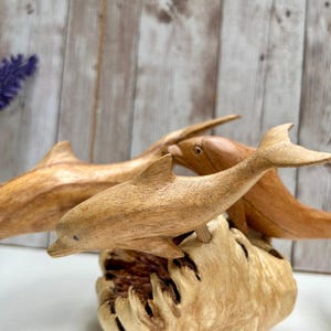 Peut inclure: Trois sculptures de dauphins en bois, chacune avec un motif de grain unique, sont expos&eacute;es sur une base en bois textur&eacute;. Les dauphins sont de diff&eacute;rentes nuances de brun, avec des nageoires et des queues sculpt&eacute;es en d&eacute;tail. L'arri&egrave;re-plan pr&eacute;sente un panneau de bois et une brindille de fleurs violettes.