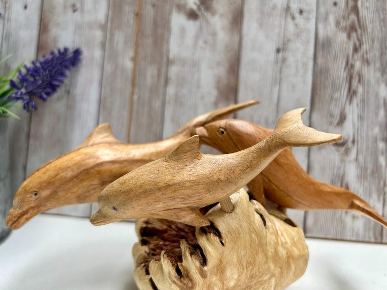 Peut inclure: Trois sculptures de dauphins en bois, chacune avec un motif de grain unique, sont expos&eacute;es sur une base en bois textur&eacute;. Les dauphins sont de diff&eacute;rentes nuances de brun, avec des nageoires et des queues sculpt&eacute;es en d&eacute;tail. L'arri&egrave;re-plan pr&eacute;sente un panneau de bois et une brindille de fleurs violettes.