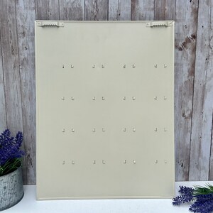 Op de afbeelding: Een witte metalen sleutelhanger met 16 haken. De sleutelhanger is gemonteerd op een houten muur.