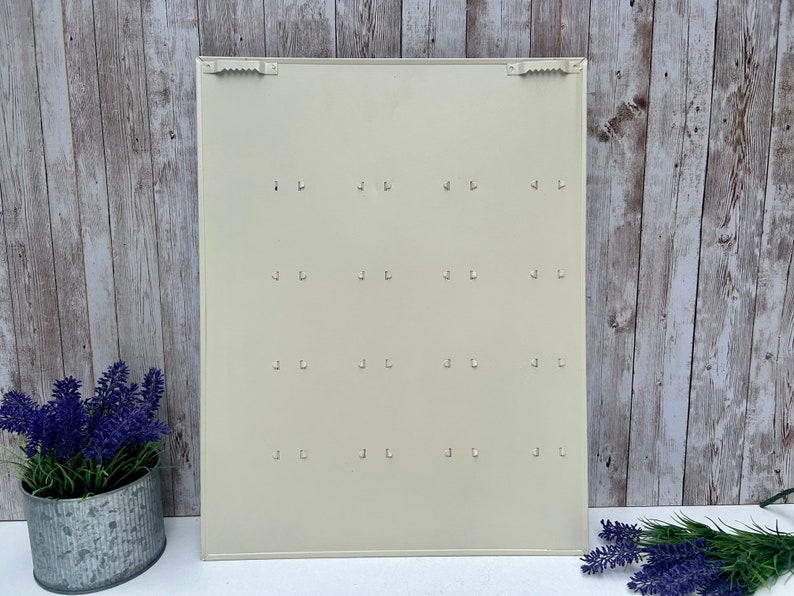 Op de afbeelding: Een witte metalen sleutelhanger met 16 haken. De sleutelhanger is gemonteerd op een houten muur.