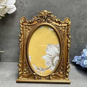 May include: An ornate gold picture frame with an oval aperture. The frame features detailed floral and scroll patterns. The background within the frame is yellow with a white floral design. The frame stands on a flat surface.