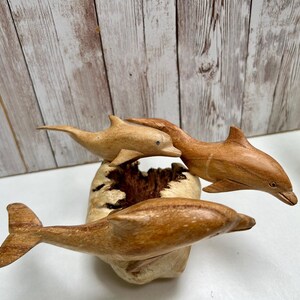 Peut inclure: Trois sculptures de dauphins en bois de diff&eacute;rentes nuances de brun sont pr&eacute;sent&eacute;es sur une base claire et textur&eacute;e. Les dauphins semblent sauter, leurs corps courb&eacute;s et leurs nageoires d&eacute;ploy&eacute;es. L'arri&egrave;re-plan pr&eacute;sente un mur en planches de bois.