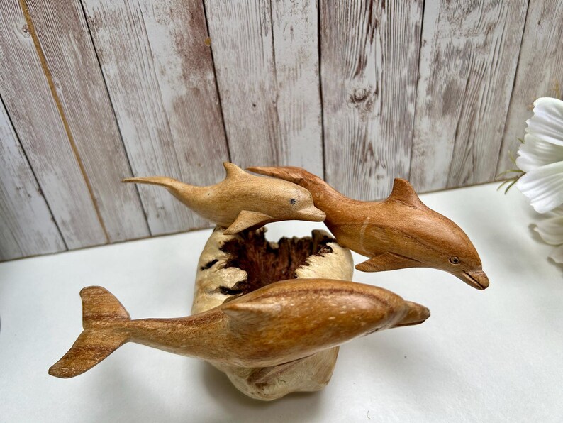 Peut inclure: Trois sculptures de dauphins en bois de diff&eacute;rentes nuances de brun sont pr&eacute;sent&eacute;es sur une base claire et textur&eacute;e. Les dauphins semblent sauter, leurs corps courb&eacute;s et leurs nageoires d&eacute;ploy&eacute;es. L'arri&egrave;re-plan pr&eacute;sente un mur en planches de bois.