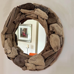 May include: Circular mirror with a frame made from driftwood. The frame is constructed from various pieces of light and dark brown wood. The mirror reflects a room with a framed picture and a candle.
