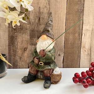 May include: A green and brown gnome figurine sitting with a fishing rod in hand. The gnome is wearing a green hat and a brown waistcoat. The gnome is holding a basket with white birds in it. The gnome is sitting on a brown rock.