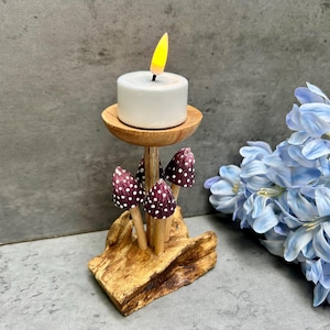 May include: A wooden candle holder with a white candle and a faux flame. The holder features decorative mushroom accents with white polka dots and is set on a piece of natural wood. Blue flowers are in the background.