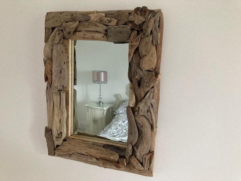 May include: A rectangular mirror with a unique frame made of driftwood. The frame is made of various pieces of driftwood, some of which are quite large. The mirror is reflecting a room with a lamp and a bed.