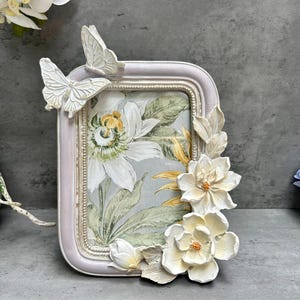 Peut inclure: Un cadre photo rectangulaire avec un motif floral. Le cadre est violet clair avec des papillons et des fleurs blancs. L'image à l'intérieur du cadre a un motif floral avec des fleurs blanches et jaunes.